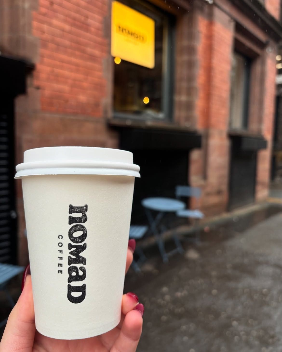 A white nomad takeaway cup held up on a wet Northern Quarter street, red-brick warehouses behind